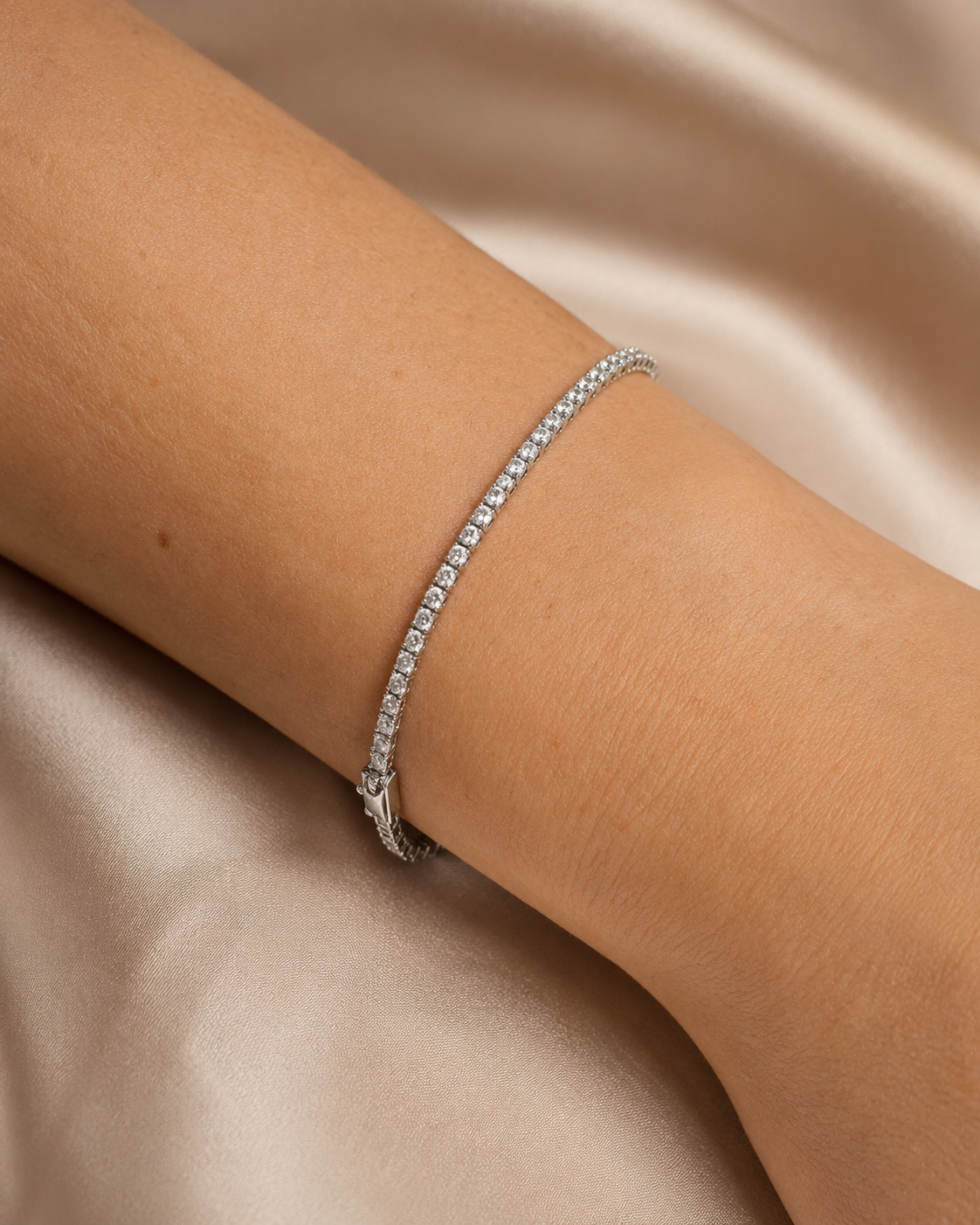 A delicate silver tennis bracelet with small clear stones, shown worn on a person's wrist against a soft beige satin background.
