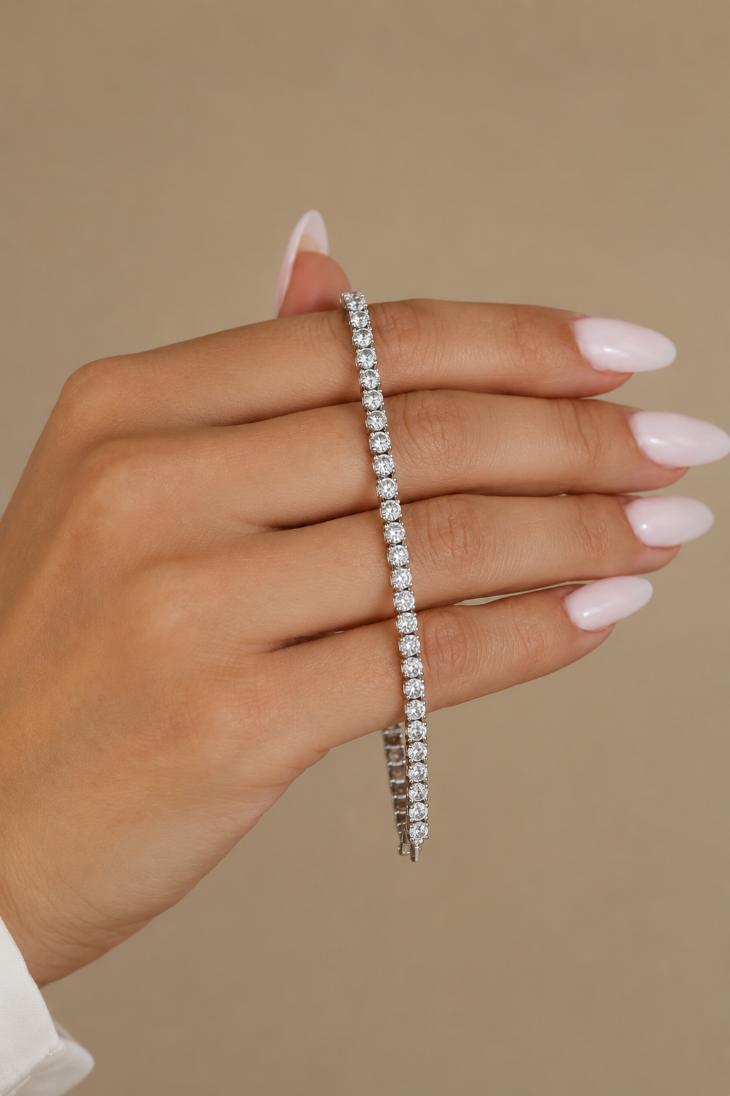A close-up of a hand with pale pink manicured nails wearing a delicate silver tennis bracelet with sparkling clear stones, set against a solid warm beige background.