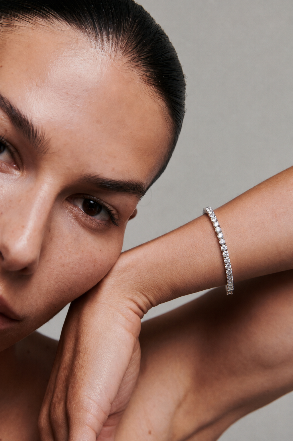 A close-up lifestyle shot of a model wearing a delicate silver tennis bracelet on her wrist, with her face and hand resting near the frame.